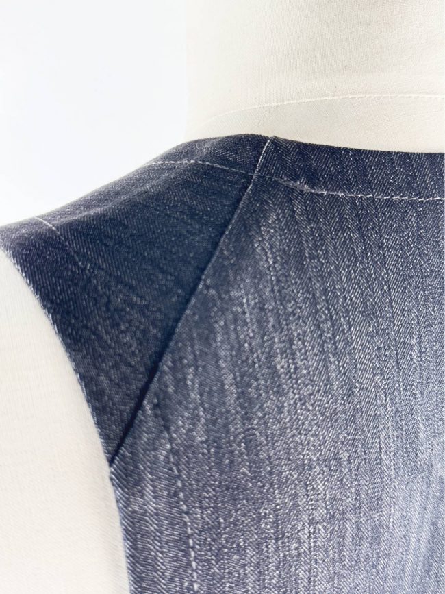 Robe trapèze sans manches en tissu effet jean noir, col en V, avec deux grandes poches plaquées. Détail couture d'épaule balancée sur les omoplates à la XVIIIè